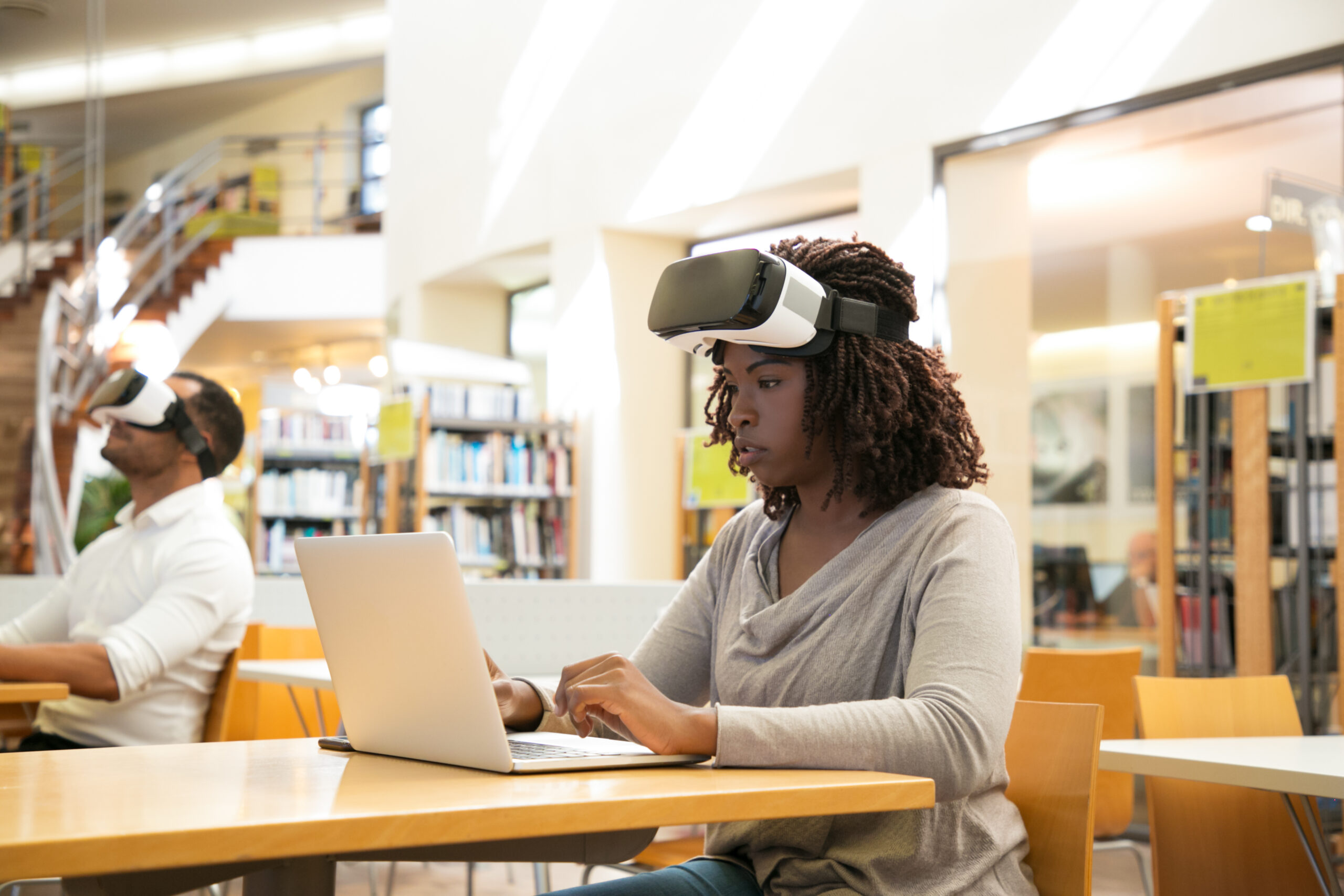 Focused black female student describing VR experience in public library. Woman with taken off virtual reality glasses, sitting at desks and using laptop. VR experience concept